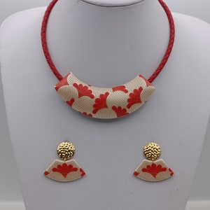 Op de afbeelding: Een rode leren choker ketting met een crèmekleurige hanger met een rood bloemenpatroon. Passende oorbellen met een crèmekleurige basis en een rood bloemenpatroon.