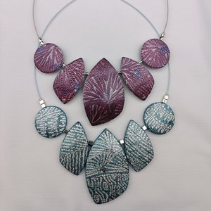 Puede incluir: Un collar de cadena plateada con dos capas de colgantes. La capa superior tiene tres colgantes en forma de hoja de color morado y plateado y dos colgantes redondos. La capa inferior tiene tres colgantes en forma de hoja de color turquesa y plateado y dos colgantes redondos.