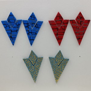 May include: Six pairs of geometric patterned earrings. Two pairs are blue with black geometric patterns, two pairs are red with black geometric patterns, and two pairs are teal with gold geometric patterns.