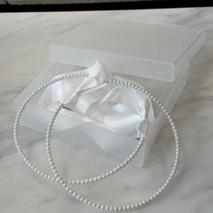 May include: Two white pearl headbands in a clear plastic box with white satin ribbon bows.