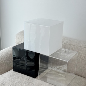 Könnte beinhalten: Drei Acrylboxen, eine weiße, eine schwarze und eine transparente, sind auf einem hellgrauen Sofa gestapelt. Die Boxen sind quadratisch und haben ein schlichtes Design.
