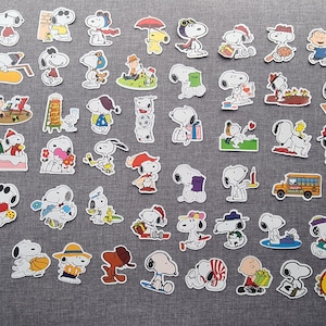 May include: A selection of Snoopy stickers, showcasing the character in diverse scenarios. The stickers depict Snoopy engaged in various activities, such as playing sports, wearing hats, and interacting with other Peanuts characters. The stickers are colourful with white borders.