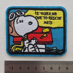 May include: An embroidered patch featuring a cartoon dog wearing goggles and a red jacket. The dog is holding a yellow sign that says "Is there no one to rescue me?" The patch is on a blue background.