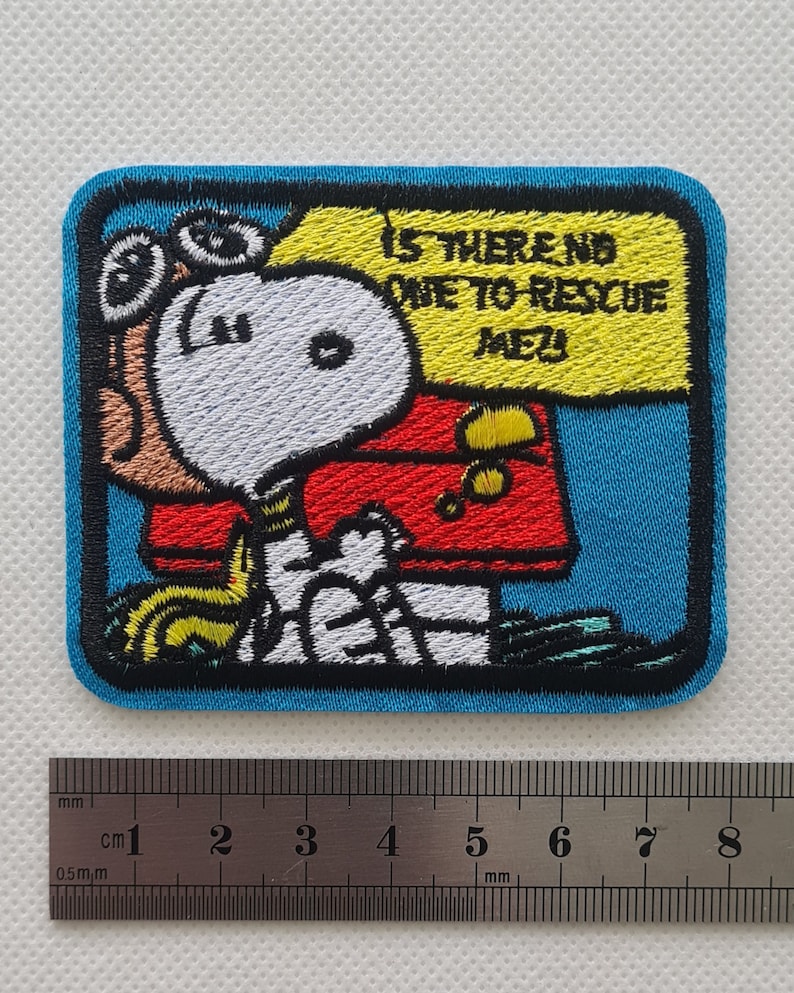 May include: An embroidered patch featuring a cartoon dog wearing goggles and a red jacket. The dog is holding a yellow sign that says "Is there no one to rescue me?" The patch is on a blue background.