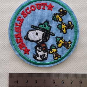 May include: A blue and green embroidered patch with a cartoon dog wearing a ranger hat and the text "Eagle Scout" in red. There are three yellow and green cartoon birds on the patch.