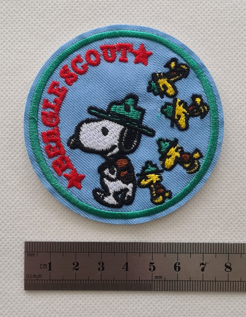May include: A blue and green embroidered patch with a cartoon dog wearing a ranger hat and the text "Eagle Scout" in red. There are three yellow and green cartoon birds on the patch.