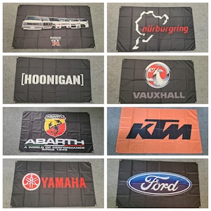 May include: A collection of eight rectangular fabric banners featuring various automotive brand logos and designs, including Nissan GT-R, Nürburgring, Hoonigan, Vauxhall, Abarth, KTM, Yamaha, and Ford, each with metal grommets in the corners.
