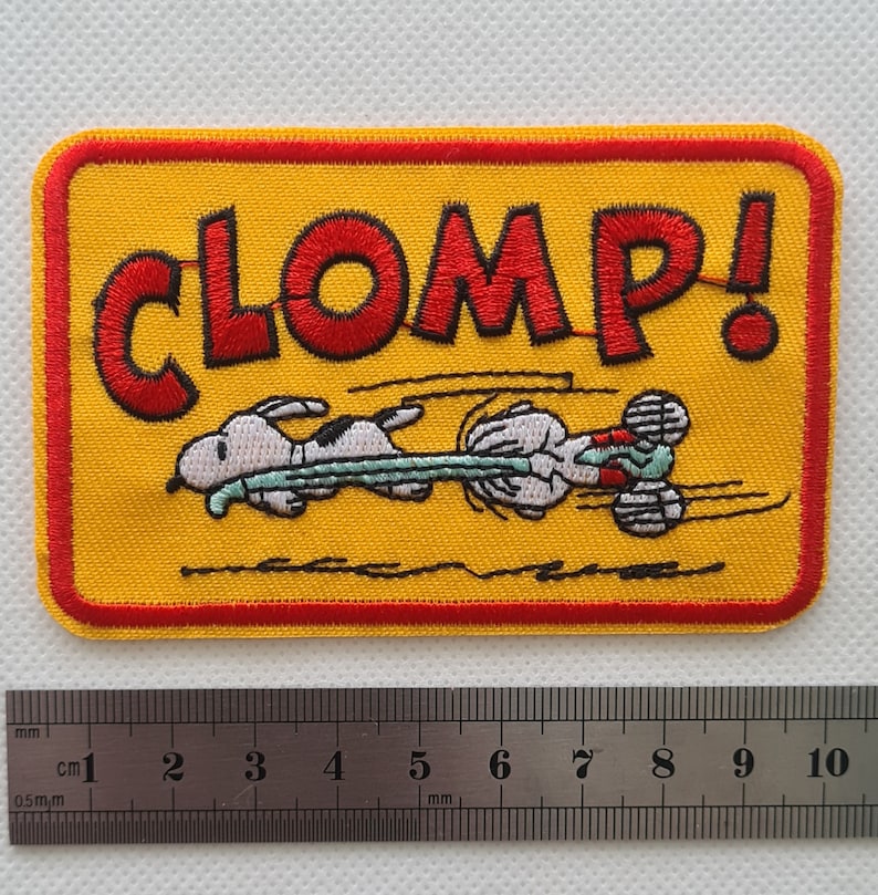 May include: A yellow embroidered patch with a red border. The patch features a cartoon dog running with the word "CLOMP!" in red letters.