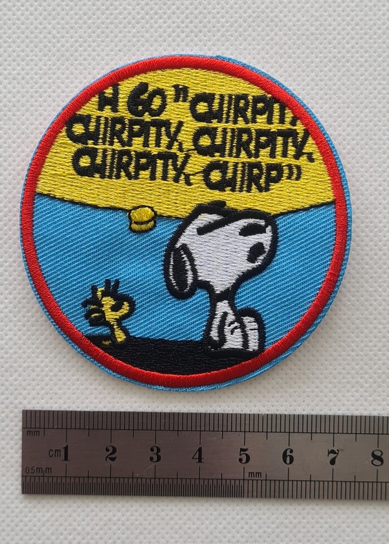May include: A round, blue and yellow embroidered patch featuring Snoopy and Woodstock. The text on the patch reads "A60' CHIRPO CHIRPITY, CHIRPITY, CHIRPITY CHIRP".