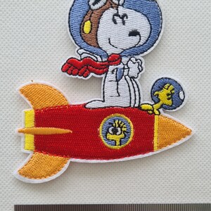 May include: Embroidered iron-on patch featuring Snoopy in a blue helmet and goggles, flying a red rocket with yellow flames. Snoopy is wearing a red scarf and holding a yellow bird.