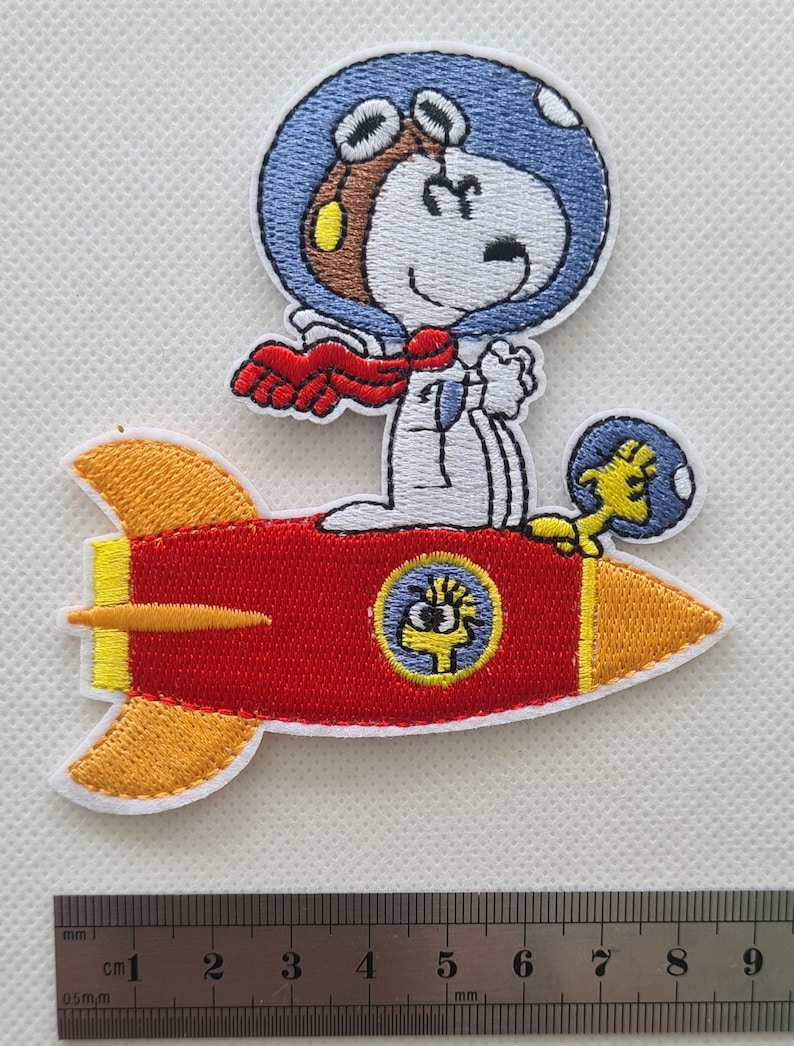 May include: Embroidered iron-on patch featuring Snoopy in a blue helmet and goggles, flying a red rocket with yellow flames. Snoopy is wearing a red scarf and holding a yellow bird.