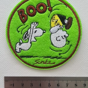 May include: A green circular embroidered patch with a brown border. The patch features a cartoon image of Snoopy and Charlie Brown. Snoopy is running and Charlie Brown is falling backwards with the word "BOO!" above them. The patch is signed "Senka" in brown thread.