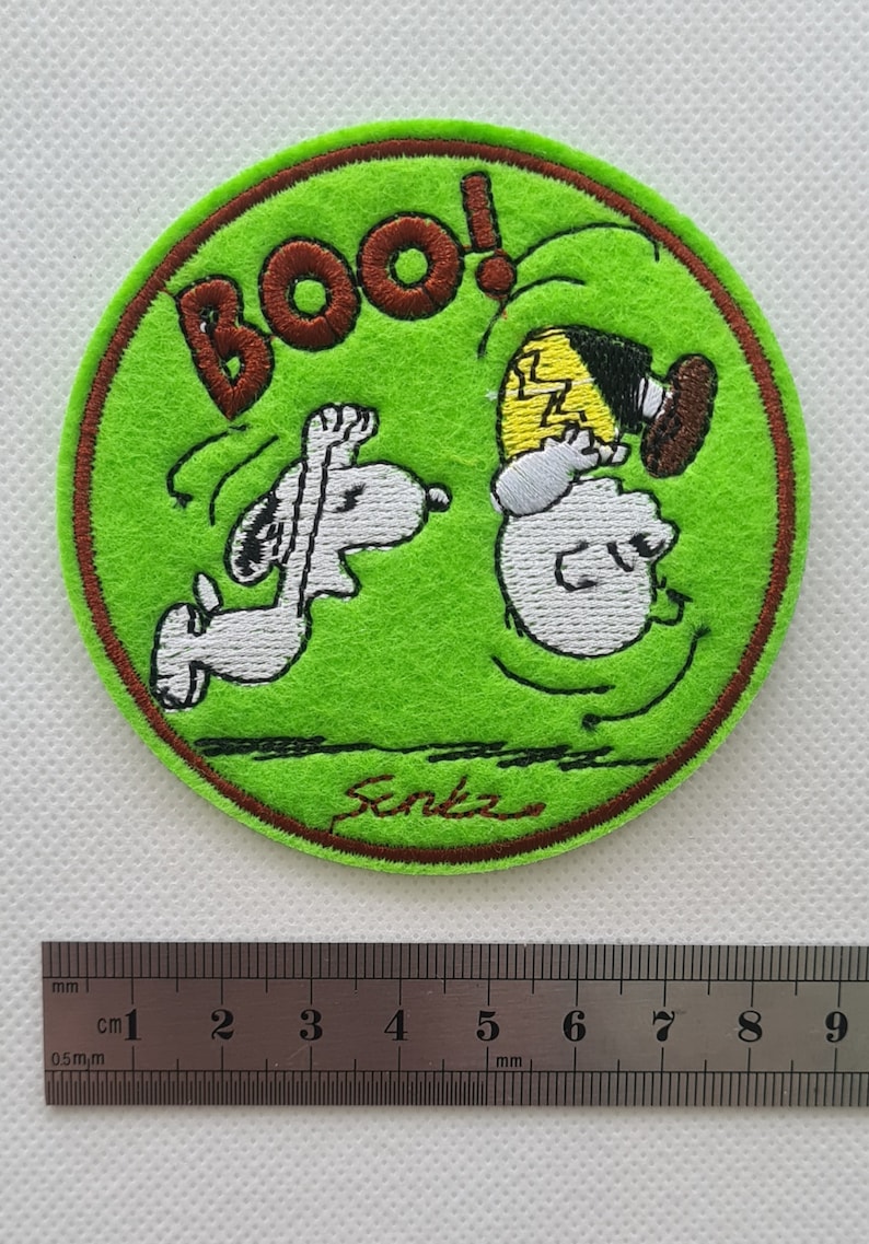 May include: A green circular embroidered patch with a brown border. The patch features a cartoon image of Snoopy and Charlie Brown. Snoopy is running and Charlie Brown is falling backwards with the word "BOO!" above them. The patch is signed "Senka" in brown thread.
