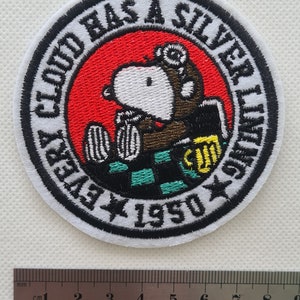 May include: A white embroidered patch with a black border. The patch features a cartoon dog wearing a pilot's helmet and holding a beer mug. The text around the dog reads "Every Cloud Has A Silver Lining 1950".