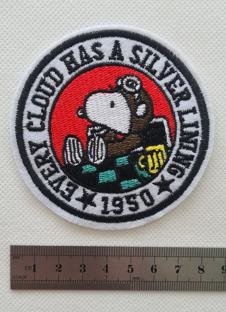 May include: A white embroidered patch with a black border. The patch features a cartoon dog wearing a pilot's helmet and holding a beer mug. The text around the dog reads "Every Cloud Has A Silver Lining 1950".