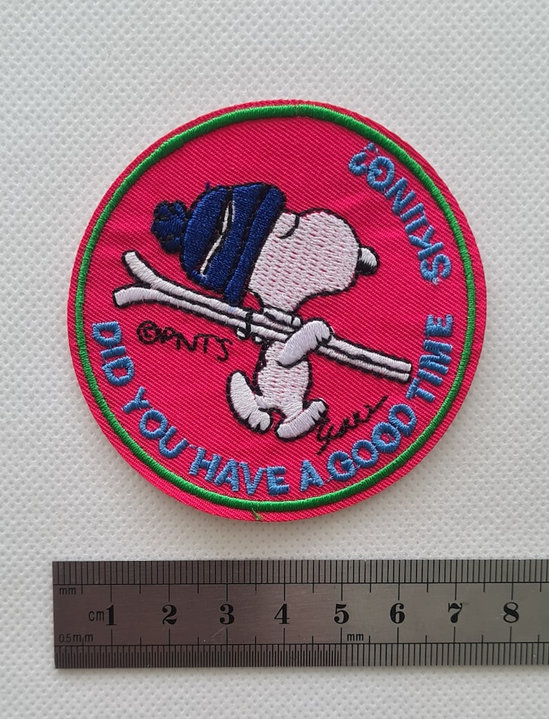May include: A pink and green embroidered patch with a cartoon dog wearing a blue hat and holding skis. The text on the patch reads "Did you have a good time skiing?"