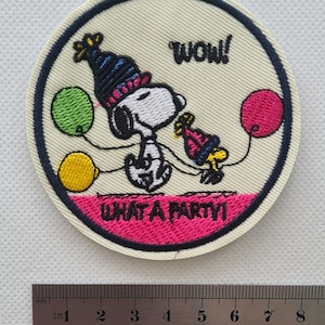 May include: A round embroidered patch featuring Snoopy wearing a party hat and holding a balloon. The patch is white with pink, blue, yellow, and green accents. The text "WOW!" and "WHAT A PARTY!" is embroidered on the patch.