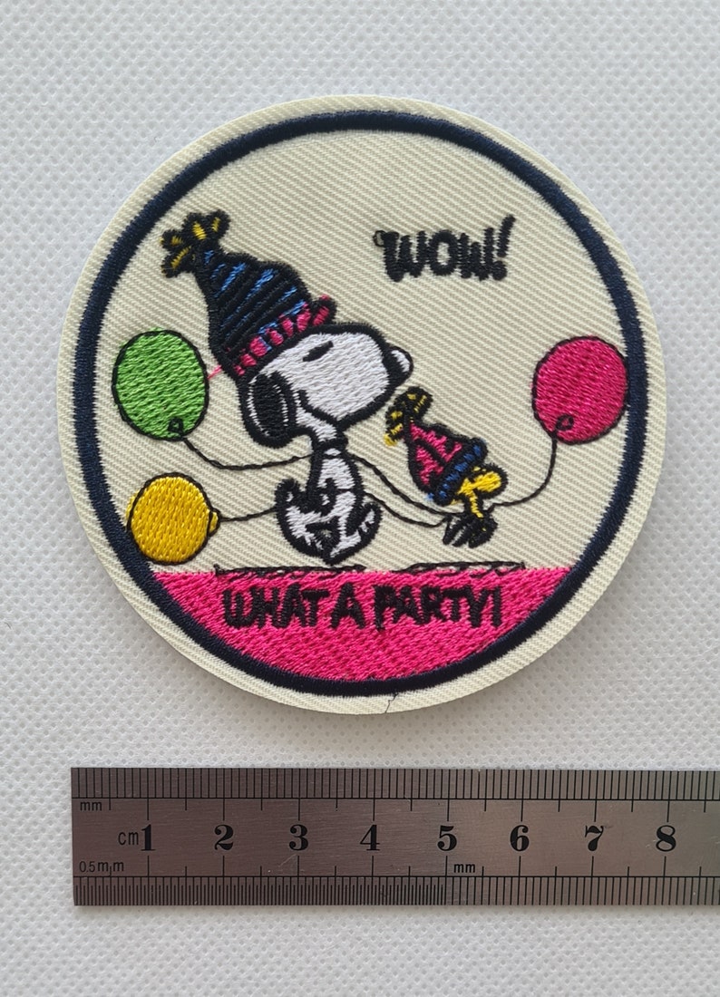 May include: A round embroidered patch featuring Snoopy wearing a party hat and holding a balloon. The patch is white with pink, blue, yellow, and green accents. The text "WOW!" and "WHAT A PARTY!" is embroidered on the patch.