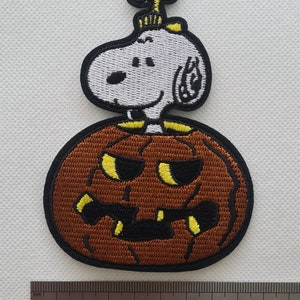 May include: An embroidered iron-on patch featuring Snoopy dressed as a pumpkin with a yellow bird on top. The pumpkin is brown with a black and yellow jack-o'-lantern face.
