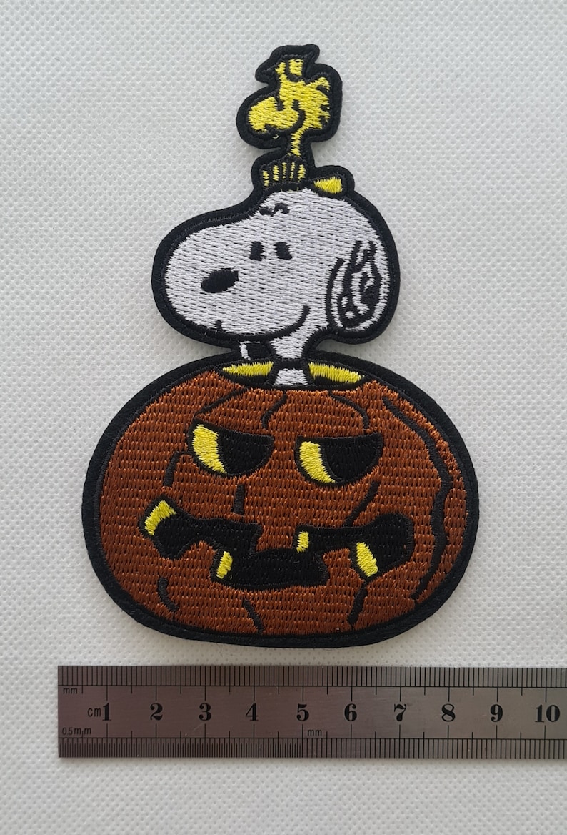 May include: An embroidered iron-on patch featuring Snoopy dressed as a pumpkin with a yellow bird on top. The pumpkin is brown with a black and yellow jack-o'-lantern face.