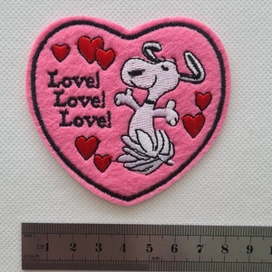 May include: Pink felt heart-shaped patch with an embroidered cartoon dog and the text "Love! Love! Love!" in pink thread. There are several red embroidered hearts around the dog.