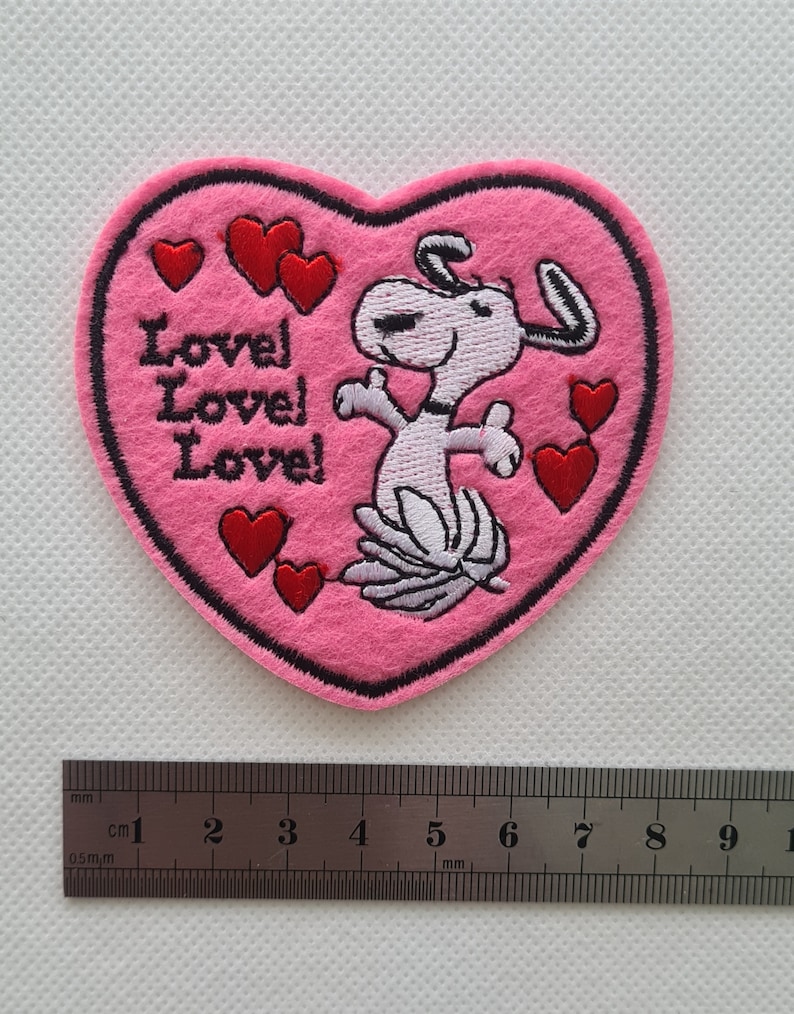 May include: Pink felt heart-shaped patch with an embroidered cartoon dog and the text "Love! Love! Love!" in pink thread. There are several red embroidered hearts around the dog.