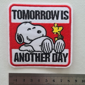 May include: White embroidered patch with a red border featuring a cartoon dog with a yellow bird. The text "TOMORROW IS ANOTHER DAY" is written in black.