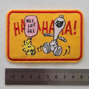 May include: Yellow embroidered patch with a red border featuring a cartoon dog with a black eye mask and a speech bubble that says "Hee Hee Hee" and the text "HAHAHA!"