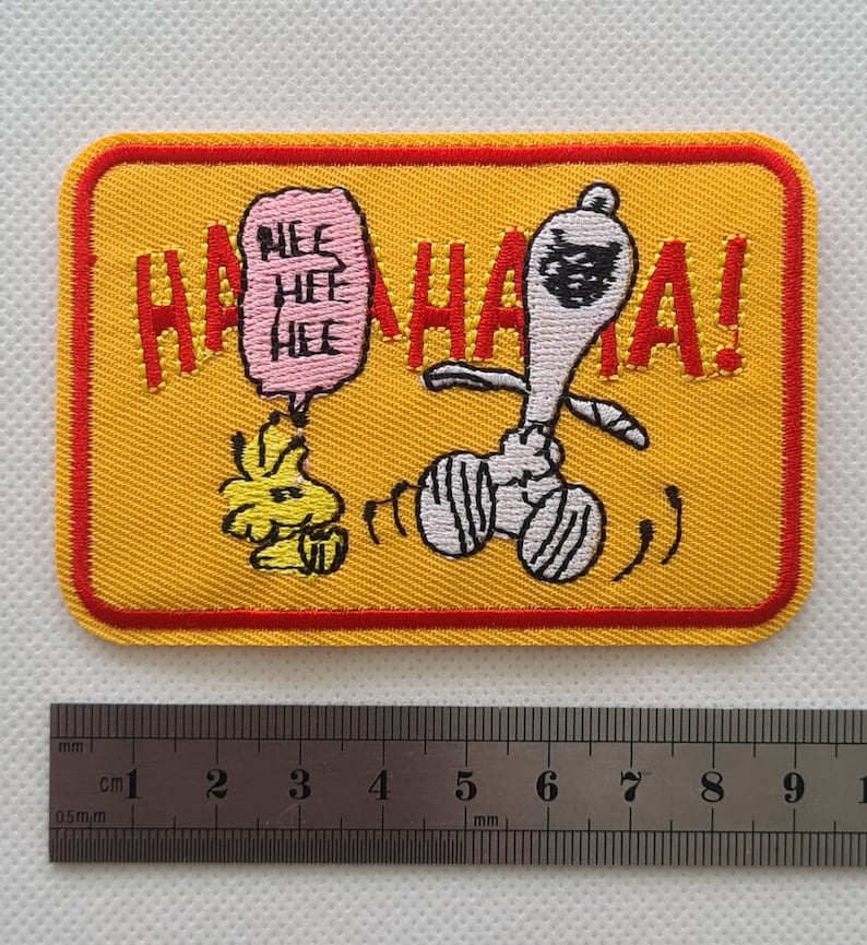 May include: Yellow embroidered patch with a red border featuring a cartoon dog with a black eye mask and a speech bubble that says "Hee Hee Hee" and the text "HAHAHA!"