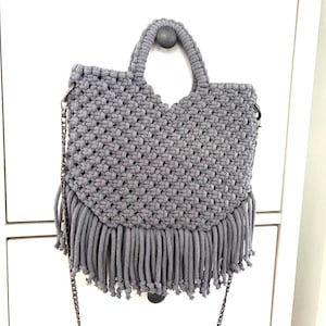 Op de afbeelding: Een grijze macramé handtas met een geweven knooppatroon, twee geïntegreerde handvatten en een lange franje langs de onderrand. De tas is voorzien van een afneembare schouderriem van donkere metaalketting aan zijringen.