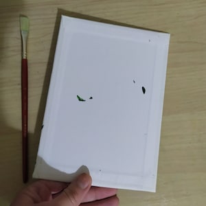 May include: A white canvas stretched over a wooden frame, held by a hand. A red-handled paintbrush with a flat, rectangular head lies to the left. Green paint flecks are visible on the canvas. Art supplies for painting.