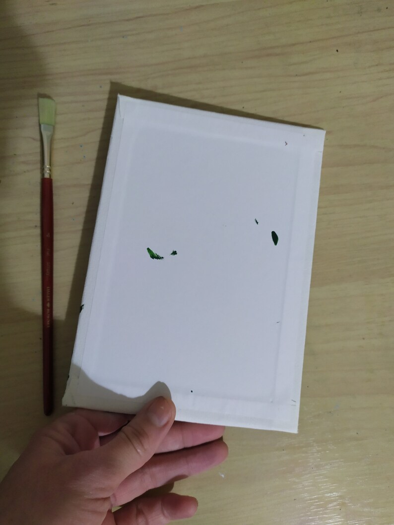 May include: A white canvas stretched over a wooden frame, held by a hand. A red-handled paintbrush with a flat, rectangular head lies to the left. Green paint flecks are visible on the canvas. Art supplies for painting.