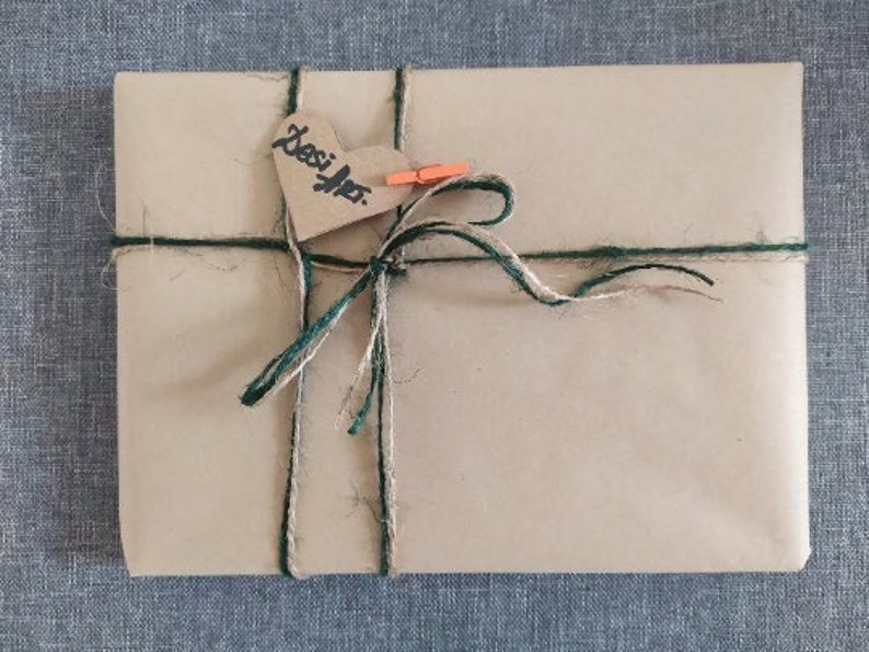 May include: A rectangular package wrapped in light brown paper, tied with twine and green string. A heart-shaped tag with the word "Desi Art" is attached with a small orange clothespin. The package is on a blue textured surface.