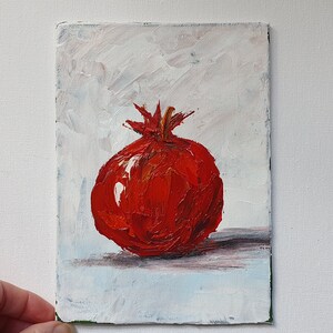 May include: Oil painting of a vibrant red pomegranate on a white and light blue background. The fruit is rendered with thick brushstrokes, showcasing its texture and color. The artwork is on a small rectangular canvas.