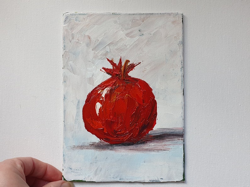 May include: Oil painting of a vibrant red pomegranate on a white and light blue background. The fruit is rendered with thick brushstrokes, showcasing its texture and color. The artwork is on a small rectangular canvas.