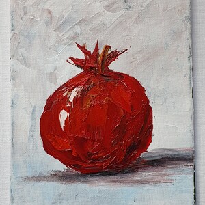 May include: A vibrant oil painting of a pomegranate. The fruit is a deep red with textured brushstrokes, set against a white and light blue background. The pomegranate's crown is a golden brown. The artwork is on a rectangular canvas.