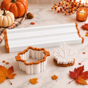 May include: A white and orange rectangular tiered display stand, a white and orange pumpkin-shaped cookie cutter, and a white maple leaf-shaped cookie stamp with an orange base. These items are arranged on a surface with scattered autumn leaves.