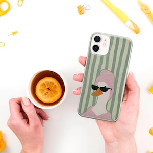 May include: A clear phone case featuring a cartoon duck wearing sunglasses and a pink towel, set against a green striped background. The phone case is held in a person's hand. A cup of tea with a lemon slice is also visible.