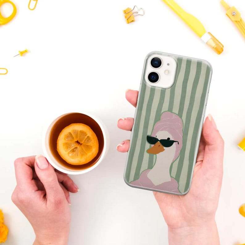 May include: A clear phone case featuring a cartoon duck wearing sunglasses and a pink towel, set against a green striped background. The phone case is held in a person's hand. A cup of tea with a lemon slice is also visible.