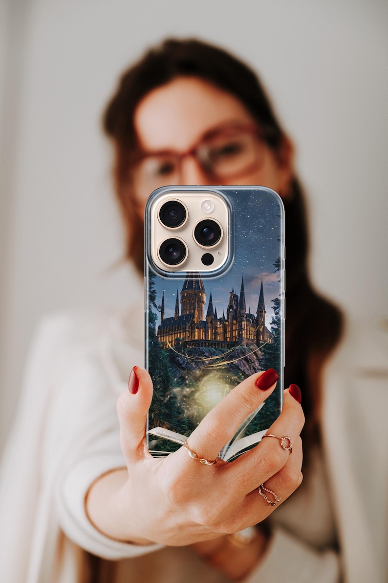 Phone case artwork showing a magical fantasy castle emerging from an open book placed on rocky ground. A soft golden light flows from the pages, illuminating the castle towers and surrounding mist. The scene is set at night with deep blue tones, glowing stars, and a dreamy atmosphere. The illustration combines elements of fantasy literature, imagination, and magic, creating a storybook-inspired design that feels warm, enchanting, and mystical.
