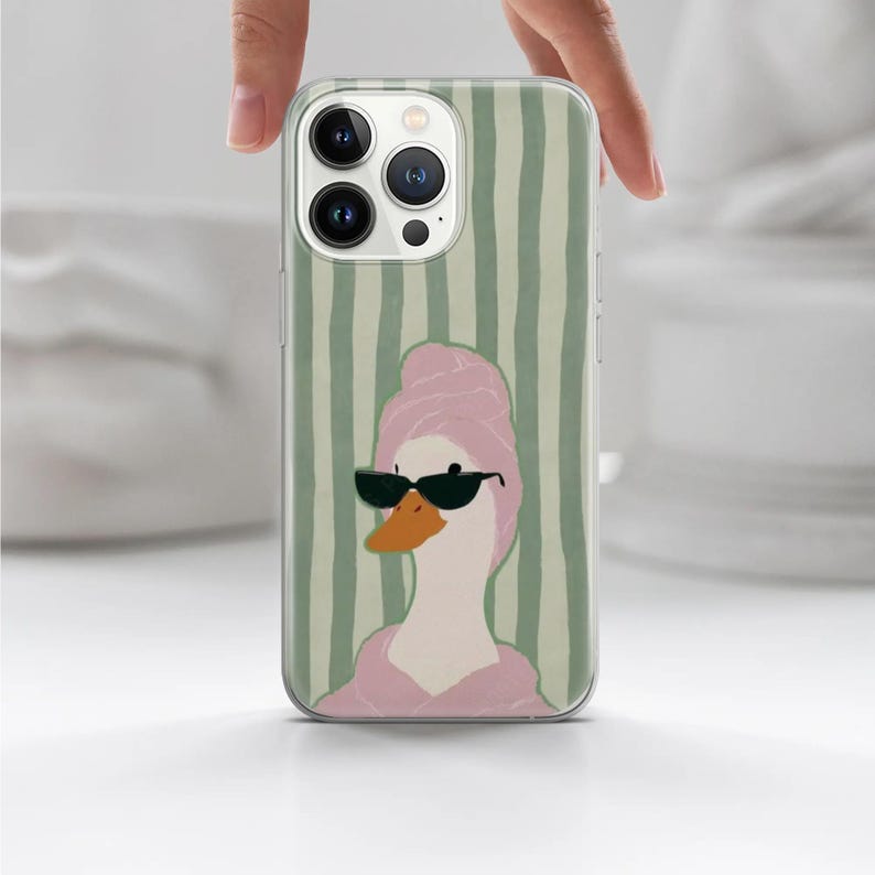 May include: A phone case featuring a cartoon duck wearing sunglasses and a pink towel, set against a background of vertical green stripes. The duck has an orange beak and is wearing black sunglasses.