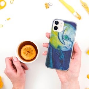 May include: A clear phone case with a colorful painting of a person playing a musical instrument in front of a crescent moon. The painting is in shades of blue, yellow, and green.