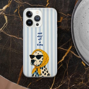 May include: A phone case with a blue and white striped background. The case features a Dalmatian dog wearing sunglasses and a yellow scarf. The text "one day at a time" is written in blue script.