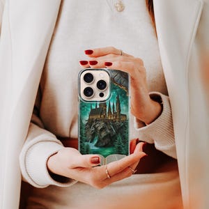 May include: A clear phone case featuring a detailed illustration of a castle on a rocky island, surrounded by a forest and a waterfall. The case has a green and teal color scheme, with a book at the bottom. The phone case is held by a person.