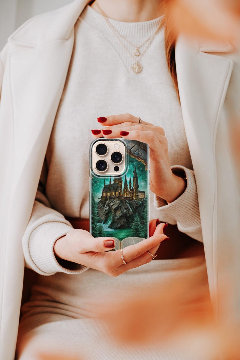 May include: A clear phone case featuring a detailed illustration of a castle on a rocky island, surrounded by a forest and a waterfall. The case has a green and teal color scheme, with a book at the bottom. The phone case is held by a person.