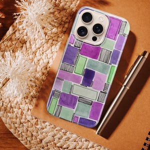 May include: A smartphone case featuring a geometric pattern in purple, green, and grey. A silver pen and a spiral-bound notebook are next to the phone case. The background includes dried flowers and a woven mat. The case has a modern design.