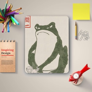 Puede incluir: Una impresión artística rectangular con una rana verde con expresión enfadada, sosteniendo un cuenco. La impresión tiene un fondo beige con un cuadrado rojo y texto japonés. Un bloc de notas muestra "Inspiring Design since 2015".