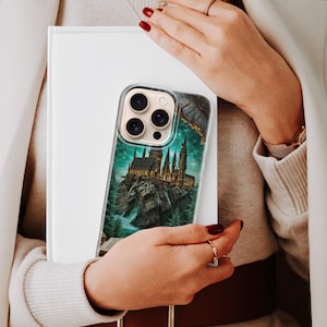 May include: Clear phone case featuring a detailed illustration of a castle on a rocky island, set against a starry night sky. The case has a transparent border and is held in front of a white book.
