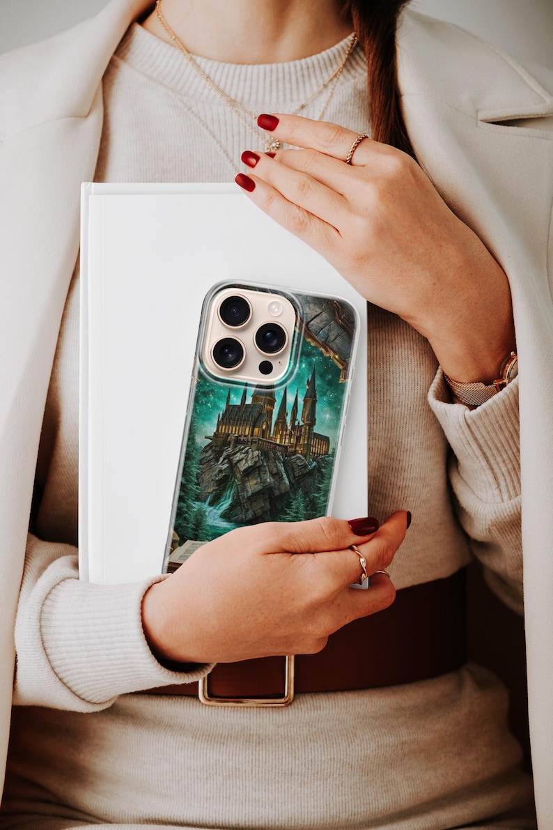 May include: Clear phone case featuring a detailed illustration of a castle on a rocky island, set against a starry night sky. The case has a transparent border and is held in front of a white book.
