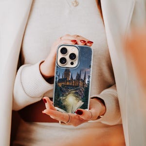 May include: A clear phone case featuring a nighttime castle scene with a starry sky. The case is held in front of a person wearing a cream-colored sweater and a white coat. The phone case has a camera and flash cutout.